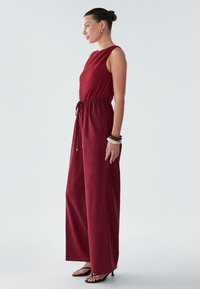 Combinaison marron avec un buste texturé, taille élastique et jambes larges. Équipée d'un détail à cordon et d'un tissu lisse. Accessoirisée avec des sandales noires.