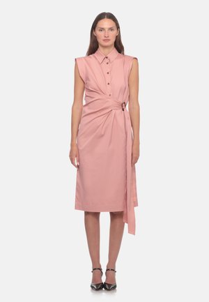 Donna che indossa un abito midi rosa senza maniche con spalline imbottite, bottoni sulla parte anteriore e dettaglio con cintura laterale, abbinato a tacchi neri a punta.