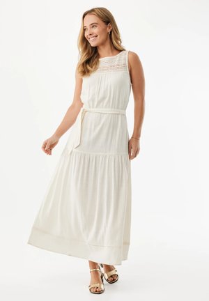 Lachende vrouw in een mouwloze witte maxi-jurk met kanten inzetstuk, ceintuur in de taille en gouden sandalen, staand tegen een witte achtergrond.