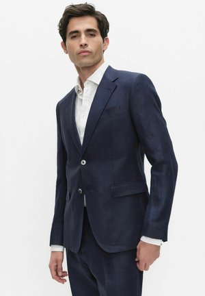 Man wearing a fitted dark blue suit with two buttons and a white dress shirt, standing against a plain light background.