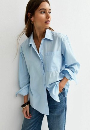 Młoda kobieta pozująca w jasnoniebieskiej, luźnej koszuli zapinanej na guziki z podwiniętymi rękawami i niebieskich dżinsach na tle białej ściany.