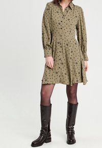 Robe boutonnée vert olive avec imprimé floral noir, manches bouffantes et jupe évasée. Associée à des bottes en cuir noir montantes jusqu'aux genoux.