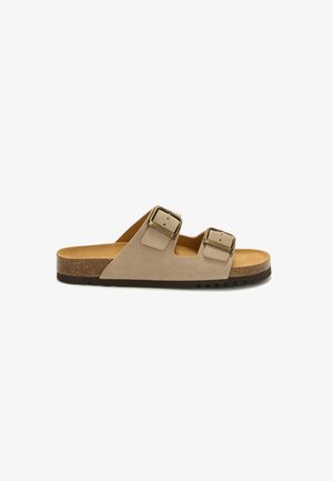 Beżowy zamszowy sandał na płaskiej podeszwie ze dwoma regulowanymi metalowymi klamrami, korkową wkładką i czarną teksturowaną gumową podeszwą.