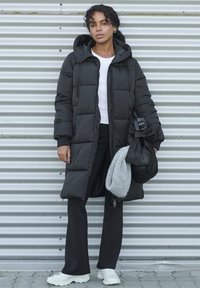 Black puffer coat with a hood, featuring a quilted design. Paired with a white shirt and black pants. Holds a gray scarf and a bag.