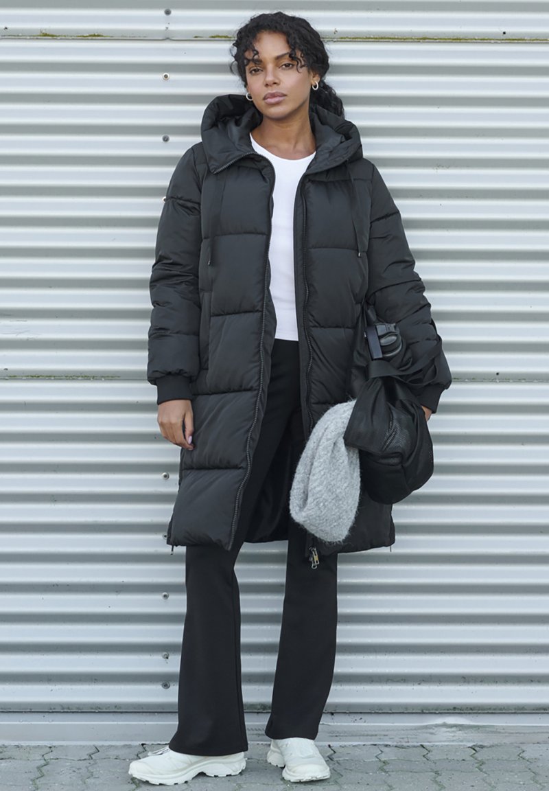 Black puffer coat with a hood, featuring a quilted design. Paired with a white shirt and black pants. Holds a gray scarf and a bag.