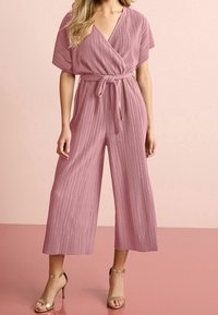 Mujer que lleva un mono plisado de color rosa empolvado con mangas cortas, escote cruzado, cintura ajustada, pantalones de pierna ancha y sandalias de tacón alto doradas.