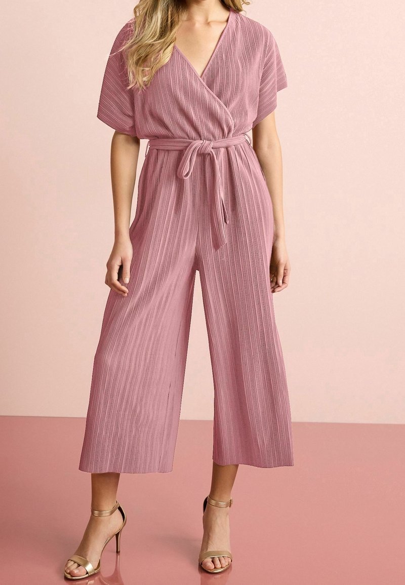 Mujer que lleva un mono plisado de color rosa empolvado con mangas cortas, escote cruzado, cintura ajustada, pantalones de pierna ancha y sandalias de tacón alto doradas.