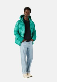 Giacca a vento verde con una texture lucida e trapuntata e cappuccio, abbinata a una maglietta nera e jeans azzurri chiari, completata da sneakers beige.