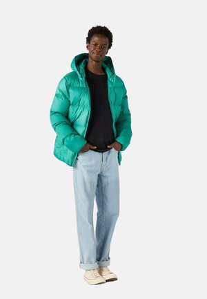 Giacca a vento verde con una texture lucida e trapuntata e cappuccio, abbinata a una maglietta nera e jeans azzurri chiari, completata da sneakers beige.