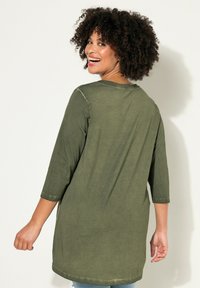 Túnica de color verde oliva, de manga larga, con escote redondeado, corte holgado y textura suave. Detalles cosidos a lo largo de las costuras.