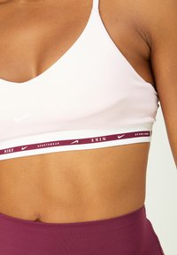 Sujetador deportivo de color rosa claro hecho de una tela elástica, con tirantes finos y una banda blanca que lleva el logo de Nike en color marrón. Textura suave con costuras mínimas.
