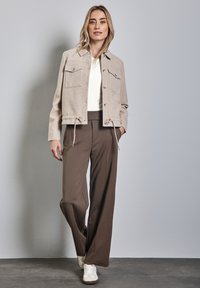 Beige cropped jakke med brystlommer, snøre i kanten, båret over en hvid top og brune vide bukser, parret med hvide sneakers.