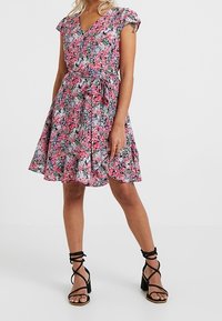 Abito floreale in tonalità di rosa e blu, con scollatura a V, maniche corte, vita aderente e gonna svasata. Abbinato a tacchi neri con cinturini.