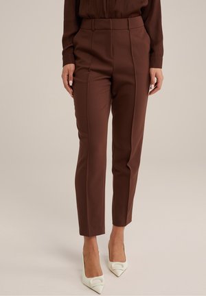 Woman wearing tailored high-waist brown trousers with front creases paired with white pointed-toe heels on a neutral background.