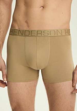 Męski tors w beżowych bokserkach z napisem "HENDERSON" na pasie, z blizną po lewej stronie brzucha.