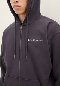 Dunkelgraue Zip-Hoodie aus Baumwollmischung. Mit einer Vordertasche, Kordelzugkapuze und weißem Logo auf der linken Brust.