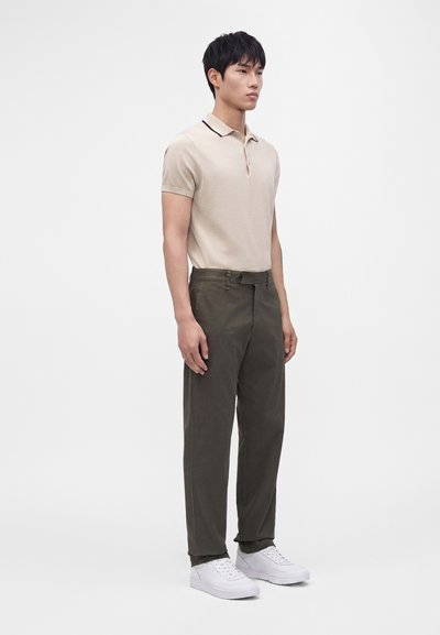 Jeune homme debout portant un polo beige à manches courtes, un pantalon vert foncé et des baskets blanches sur un fond clair uni.