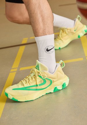 Legs wearing white Nike socks and yellow-green Nike sneakers standing on a gym floor with yellow court lines.