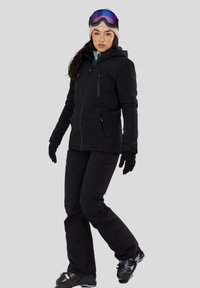 Vrouw draagt een zwarte ski-jas, broek, handschoenen, laarzen, een roze muts en skibril, staand tegen een effen achtergrond.