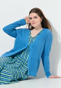 Blauer gestrickter Cardigan mit strukturierten Mustern, V-Ausschnitt und Bindebandverschluss, über einem gestreiften blau-grünen Kleid mit weitem Rock.