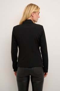 Femme aux cheveux blonds portant des boucles d'oreilles en or, un blazer noir ajusté et un pantalon en cuir noir, faisant face à un mur beige pâle.