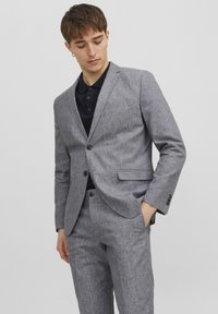 Suit grigio strutturato con vestibilità su misura, caratterizzato da un giacca monopetto e pantaloni dritti, abbinato a una polo scura.