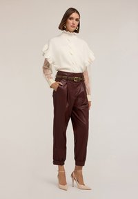 Suéter blanco con volantes, pantalones de cuero burdeos con pliegues y tacones de aguja beige. Los accesorios incluyen pendientes de aro dorados y un cinturón con tachuelas.