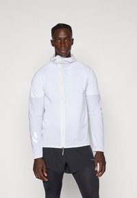 Giacca da corsa leggera bianca con cappuccio, chiusura a zip completa, dettagli riflettenti e maniche lunghe, abbinata a pantaloni corti neri.