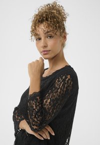 Sort blonde top med lange ærmer og bølgede kanter, intrikate blomster mønster og rund hals. Glat tekstur, tætsiddende design.