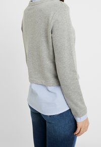 Maglione grigio corto in tessuto morbido, con maniche lunghe e scollo arrotondato, indossato sopra una camicia a righe blu e bianche.