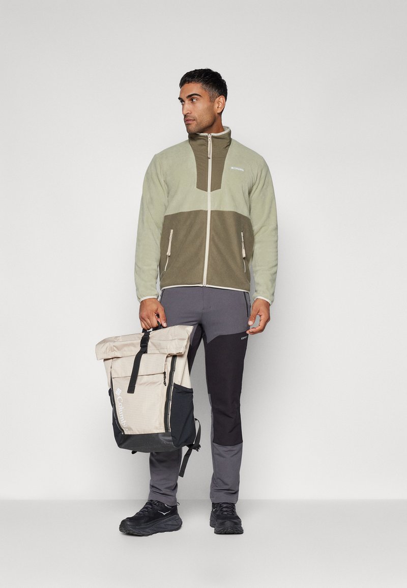Veste en polaire vert clair et olive avec fermeture éclair, associée à un pantalon gris foncé. Sac à dos à rabat marron et beige avec des bretelles.