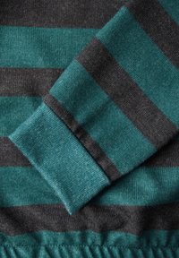 Gestreifter Stoff in Teal und Grau, mit einem strukturierten Strickdesign und einem gerippten Bündchen. Die Streifen variieren in der Breite und erzeugen einen schichtartigen Look.