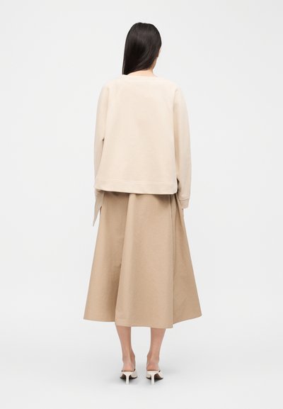 Γυναίκα με μακριά σκούρα μαλλιά που φοράει μπλούζα μπεζ με μακριά μανίκια, μίντι φούστα μπεζ και κρεμ ψηλοτάκουνα παπούτσια, στέκεται με την πλάτη της στραμμένη σε λευκό φόντο.