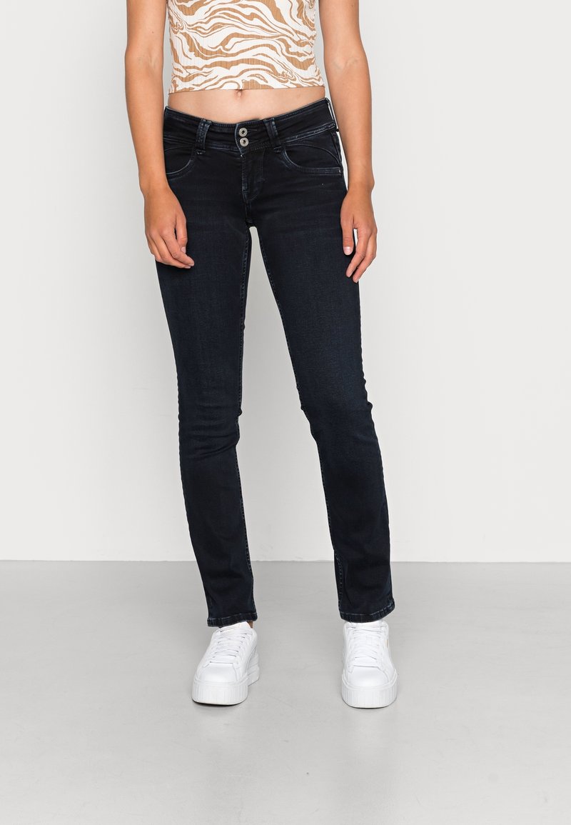 Kvinna som bär mörka skinny jeans, zebra-mönstrad crop top och vita plattformssneakers stående mot en ljus enfärgad vägg.