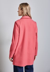 Cappotto rosa in misto lana con colletto a punta, maniche lunghe e taglio dritto. Presenta una texture liscia e arriva sotto i fianchi.