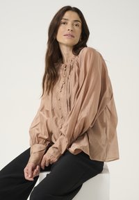 Blusa marrone chiaro con scollo arricciato, dettaglio dei bottoni e maniche lunghe a palloncino. Indossata con pantaloni neri, tessuto testurizzato, vestibilità rilassata.
