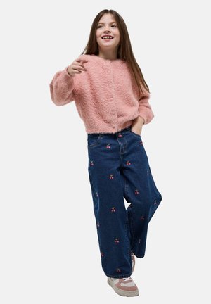 Fille aux longs cheveux bruns souriante, portant un pull rose duveteux, un jean large bleu avec une broderie de cerises et des baskets rose et blanc.