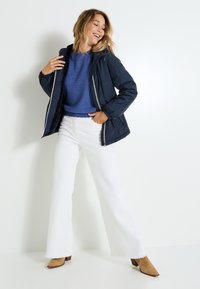 Veste rembourrée bleu marine avec fermeture éclair, portée sur un pull en laine bleu, associée à un jean blanc à jambe large et des bottes à talons beiges, se tenant devant un fond blanc.
