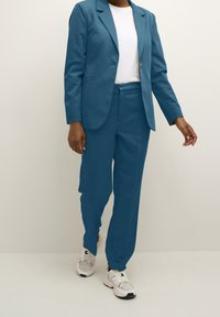 Turkusowa marynarka i dopasowane spodnie wykonane z gładkiej tkaniny, noszone na białej koszulce. Do tego białe i szare sneakersy.