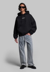 Hoodie nero oversize con tasca frontale e coulisse, abbinato a jeans a gamba larga blu chiaro e calzature nere, su uno sfondo neutro.