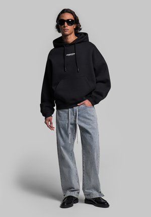 Black oversized hoodie with a front pocket and drawstring, paired with light blue wide-leg jeans and black footwear, on a neutral background.