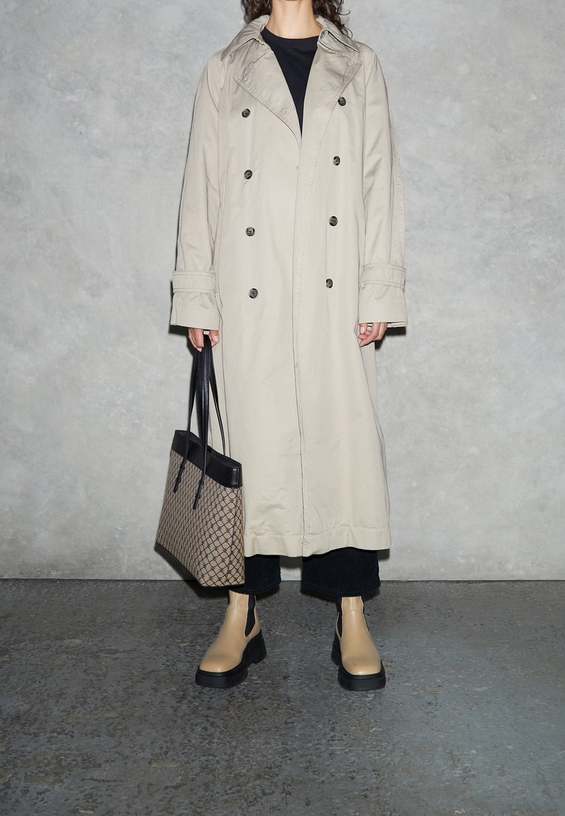 Personne portant un trench-coat beige, pantalon noir, bottes couleur sable, tenant un sac à main noir et beige à motifs devant un mur gris texturé.