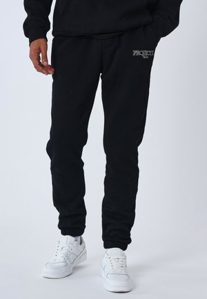 Pantalones deportivos negros con puños elásticos, con bordado "PROJECT PARIS" en el muslo. Combinados con zapatillas blancas. Material de algodón.