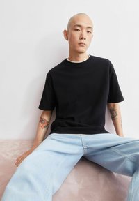 Zwart katoenen T-shirt met korte mouwen en een ronde hals. Gecombineerd met lichtblauwe spijkerbroek. Heeft een kleurrijke tatoeage op de onderarm.