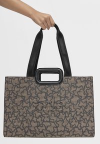 Brązowa torba na ramię w wzory o prostokątnym kształcie, z czarną rączką i kontrastowym, teksturowanym wykończeniem. Zawiera widoczny detal z logo.