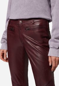 Burgunderfarbene Lederhosen zeichnen sich durch ein elegantes Design mit seitlichen Reißverschlüssen und strukturierten Einsätzen aus. Kombiniert mit einem helllilanen, langärmligen Oberteil.