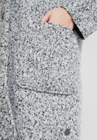 Harmaa, eläväpintainen takki, jossa on silmukalla kudottu kangaskuvio, sivutasku ja koristeellinen nappikoriste.