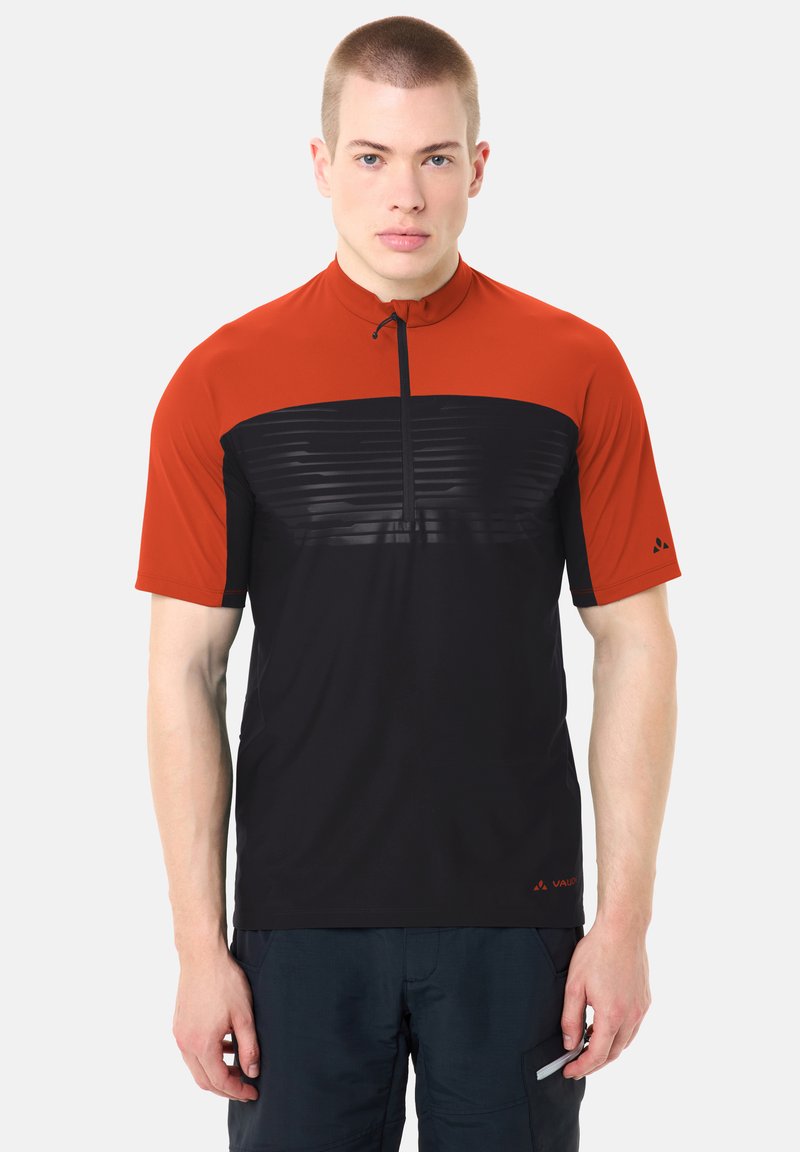 Homme portant un maillot de cyclisme à manches courtes avec une fermeture éclair courte, épaules rouges et corps noir, debout les bras détendus le long du corps.