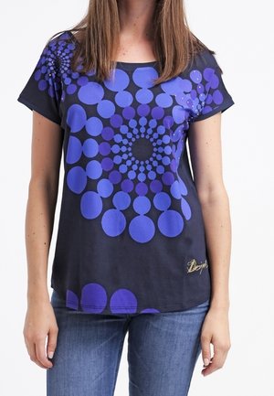Vrouw draagt een zwart T-shirt met korte mouwen met een groot spiraalpatroon van paarse cirkels, gecombineerd met een blauwe spijkerbroek.