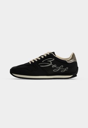 Sort lavtop sneaker med beige snørebånd og hæl, med en kursiv logo på siden og en hvid stribe langs sålen.
